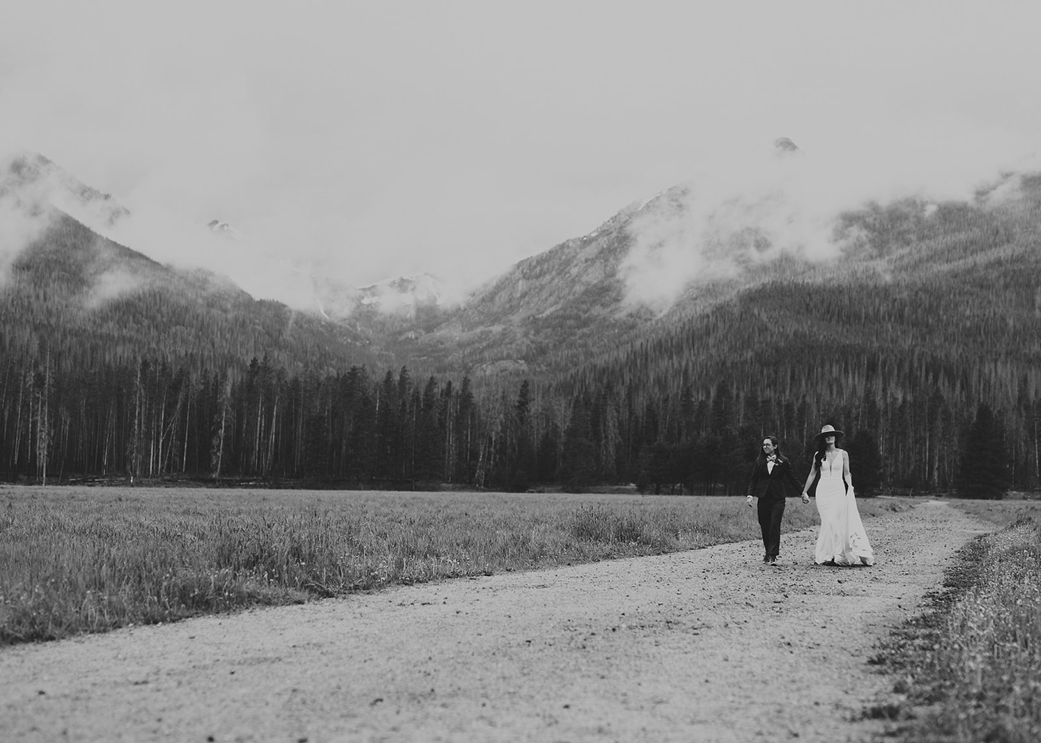 Couple holding hands and walking along dirt road in front of mountains at Colorado Airbnb wedding | McArthur Weddings and Events