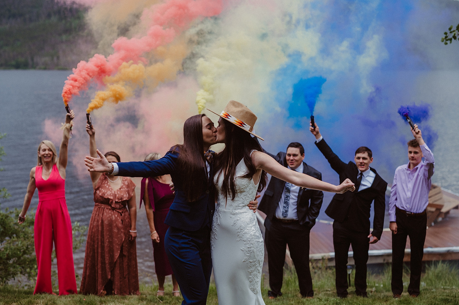 Married partners kissing each other in front of rainbow smoke bombs held by family and friends | McArthur Weddings and Events