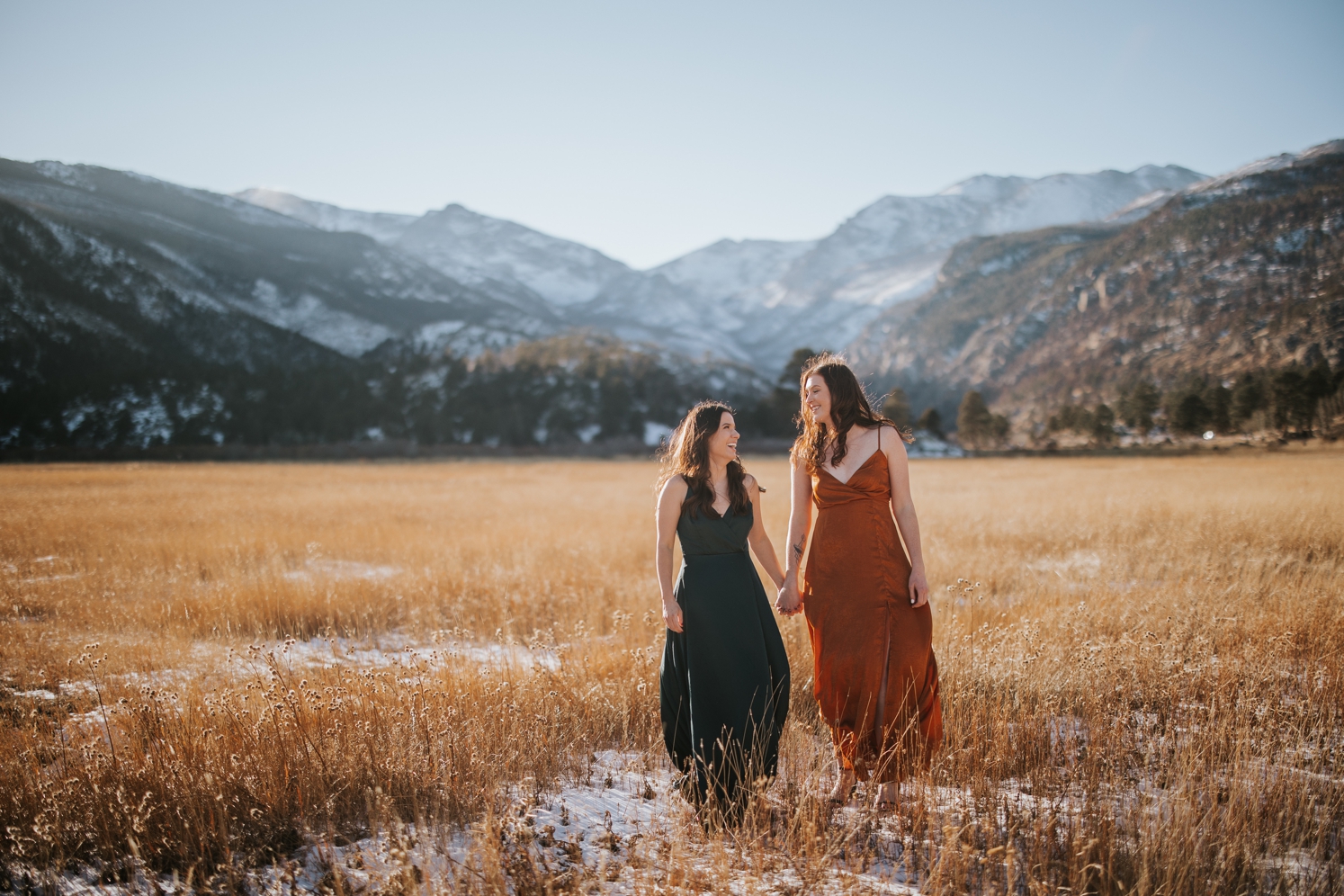 Newly engaged couple holding hands and walking through field with mountains in the background | McArthur Weddings and Events