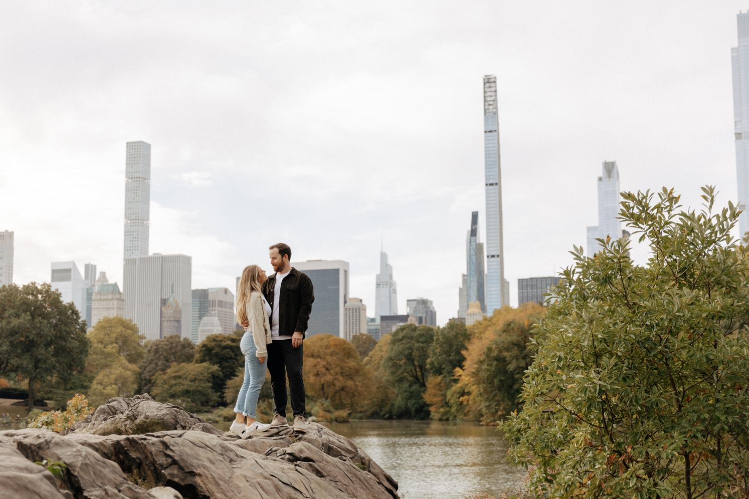 Newly engaged couple standing on rock in Central Park with NYC skyline behind them | McArthur Weddings and Events