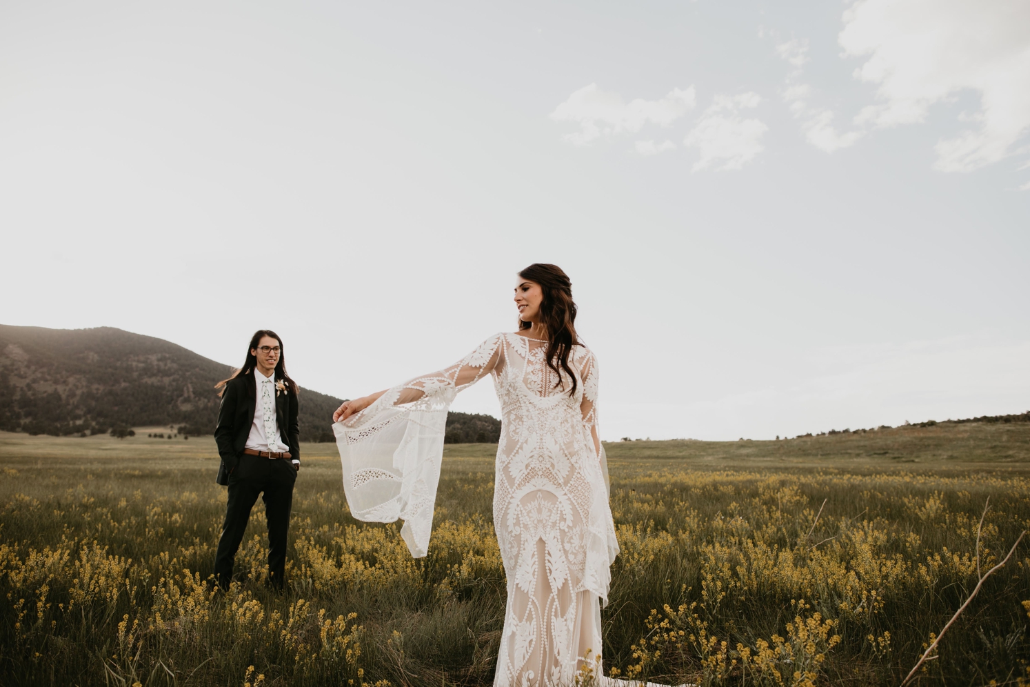 Bride wearing long sleeve lace wedding gown with bell sleeves standing in field with groom behind her | McArthur Weddings and Events