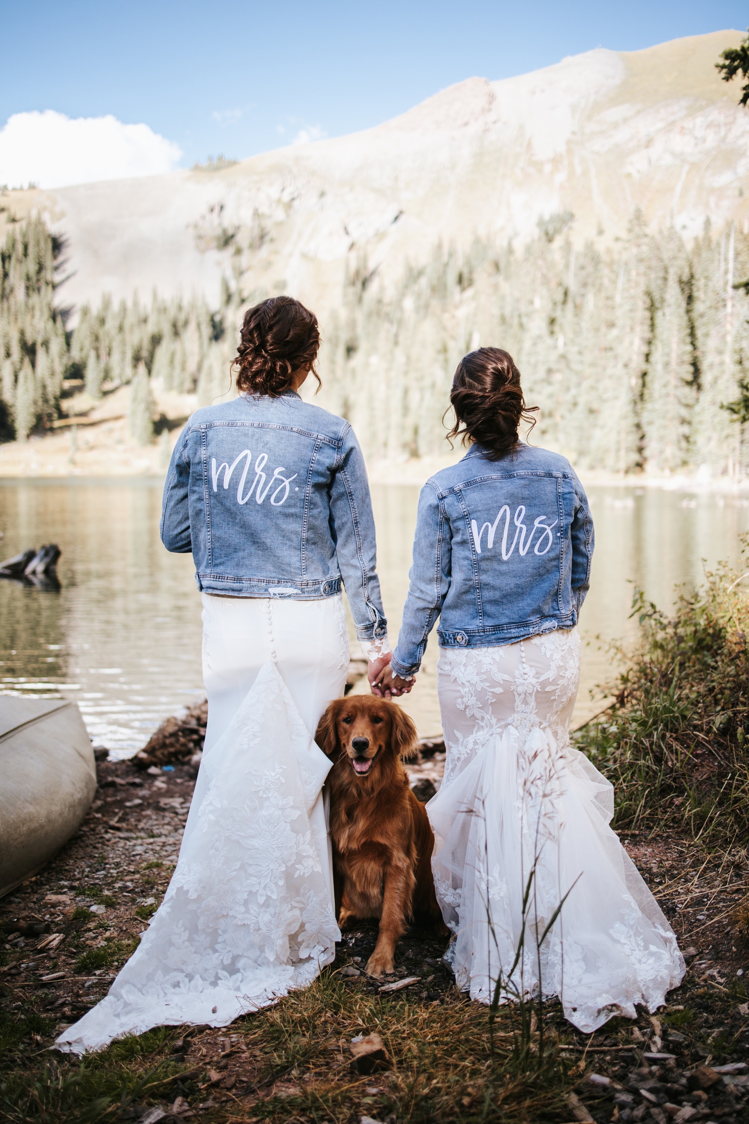 Women holding hands and wearing their wedding dresses and denim jackets with dog sitting in between them | McArthur Weddings and Events