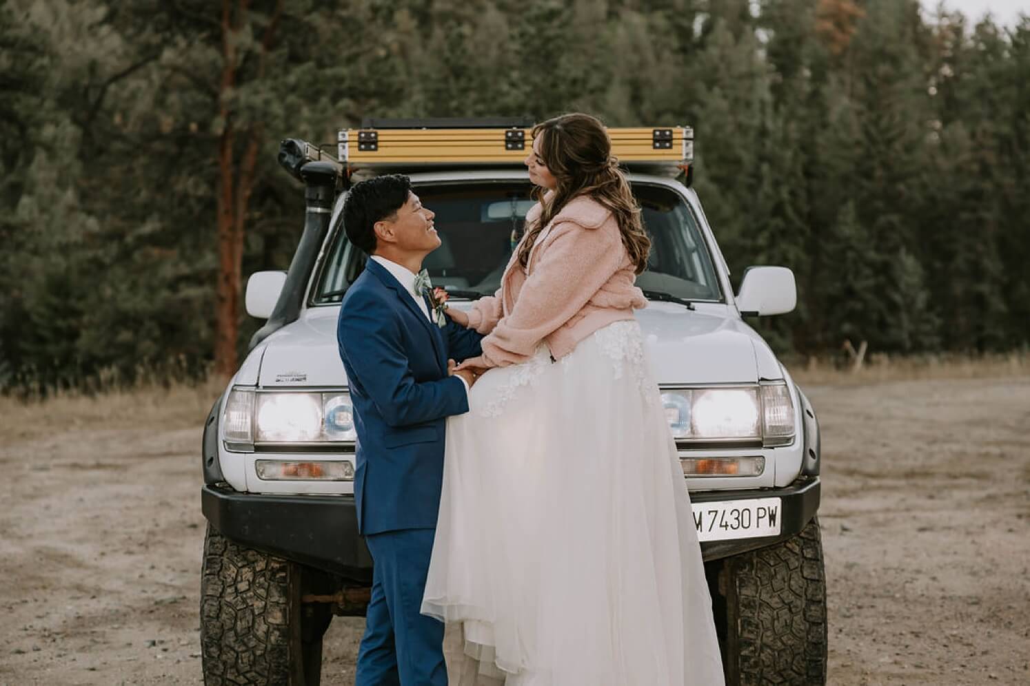Bride wearing pink jacket and sitting on top of 4Runner while groom looks at her | McArthur Weddings and Events