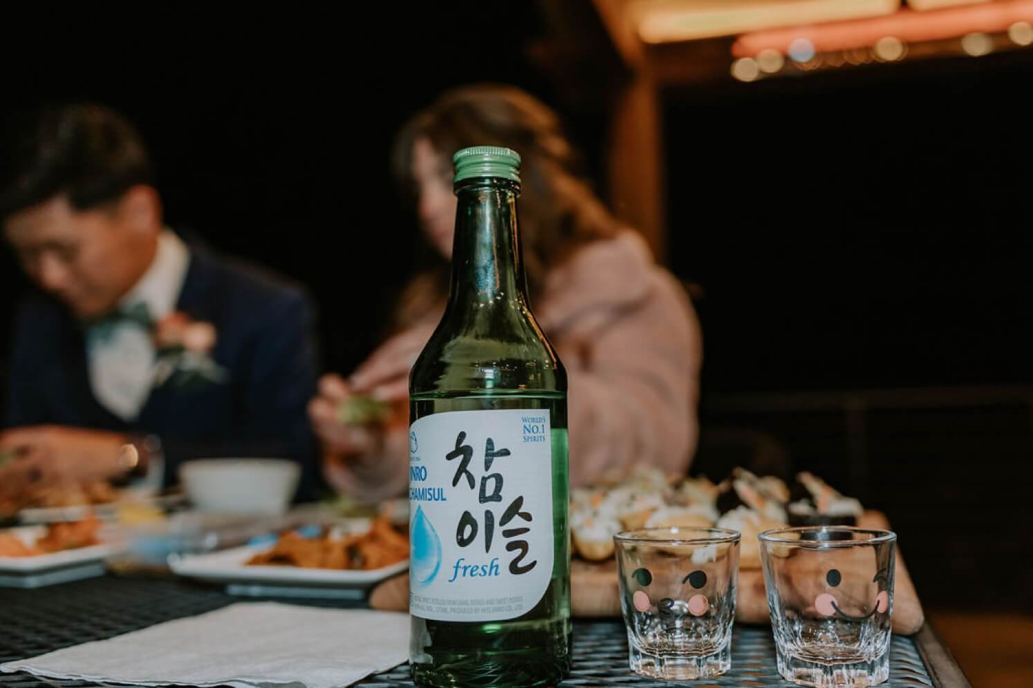 Soju and custom shot glasses in foreground with bride and groom in background | McArthur Weddings and Events