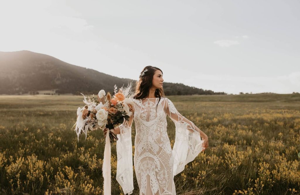 Wedding floral design: bride wearing boho wedding dress walking in field and holding boho bouquet | McArthur Weddings and Events