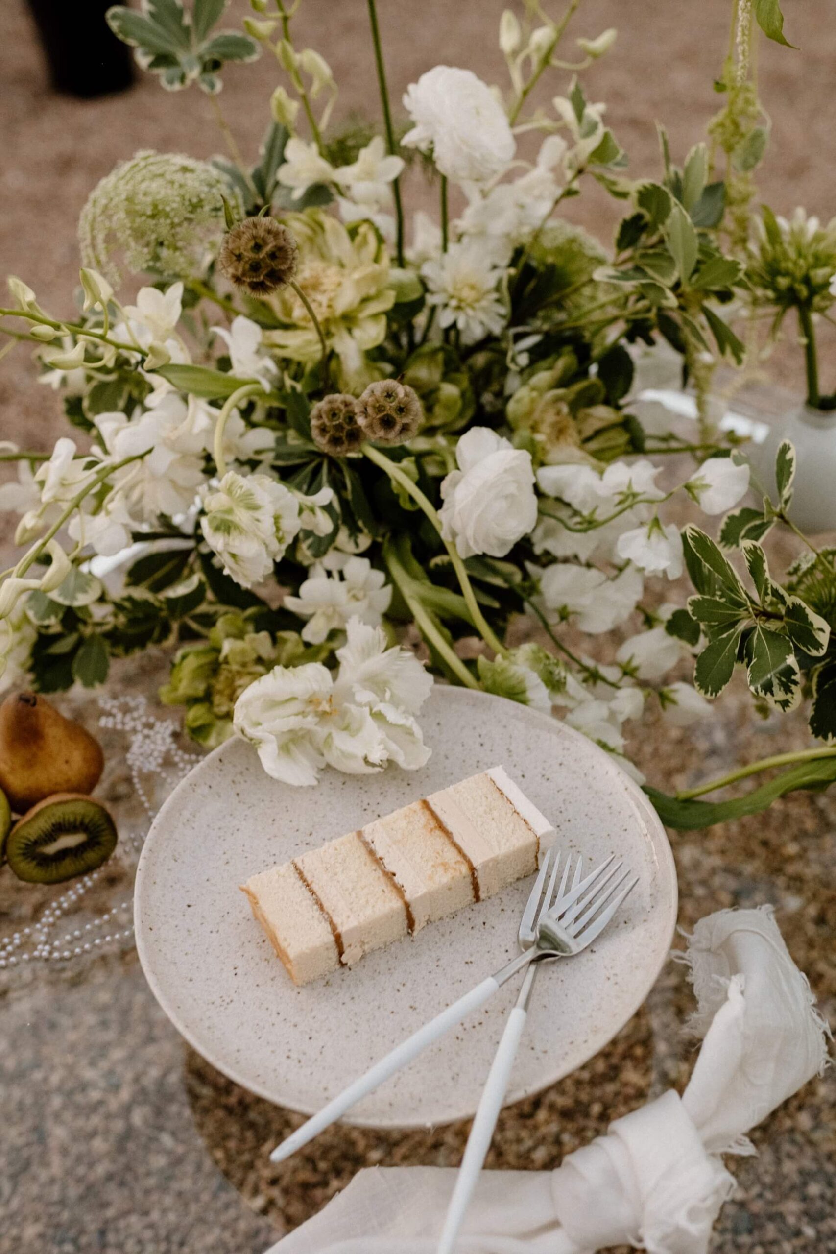 Slice of cake sitting on stone colored plate with green and white wedding flowers in the background 