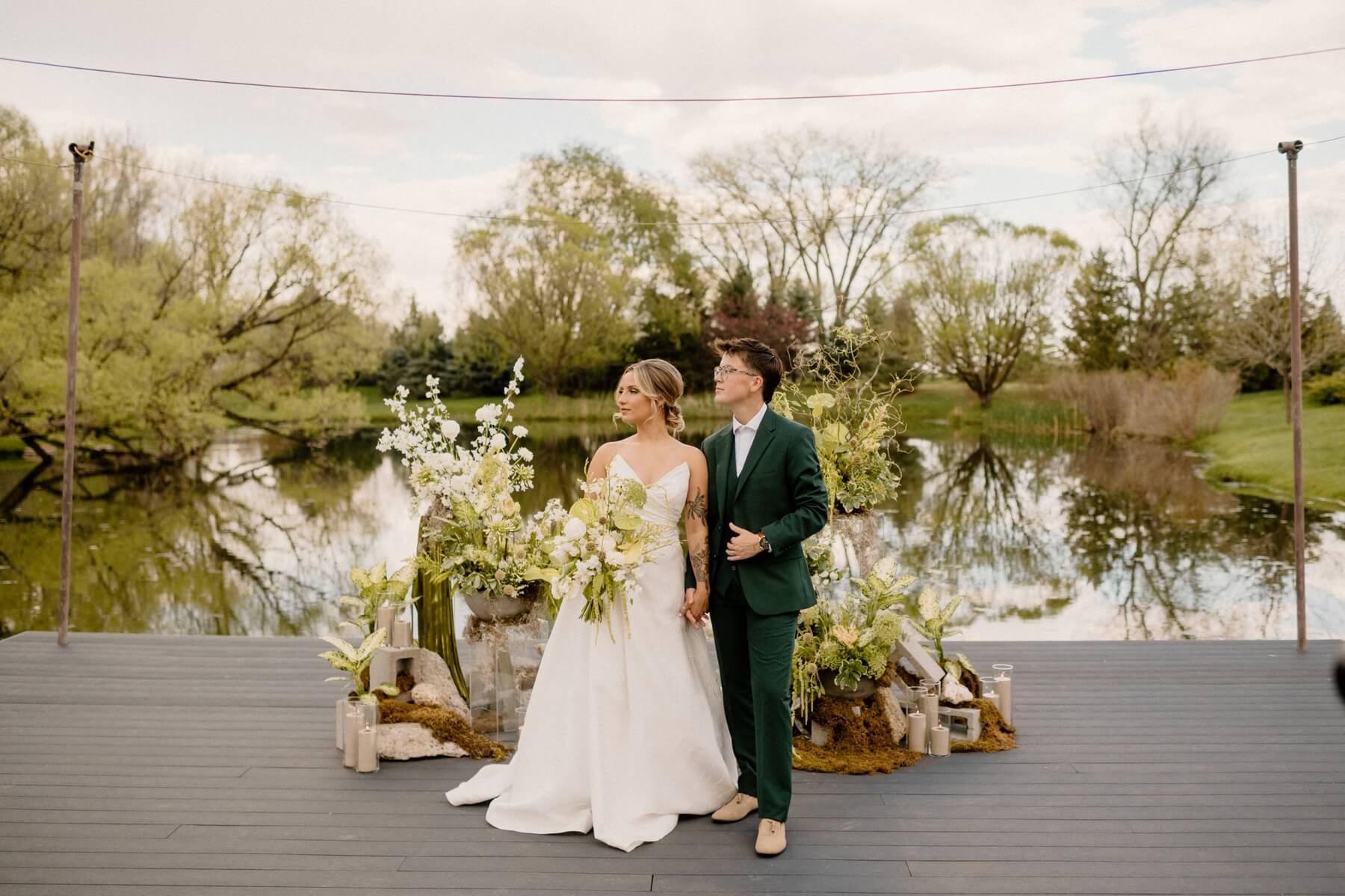 Partners holding hands and looking off into the distance in front of green and white wedding flowers