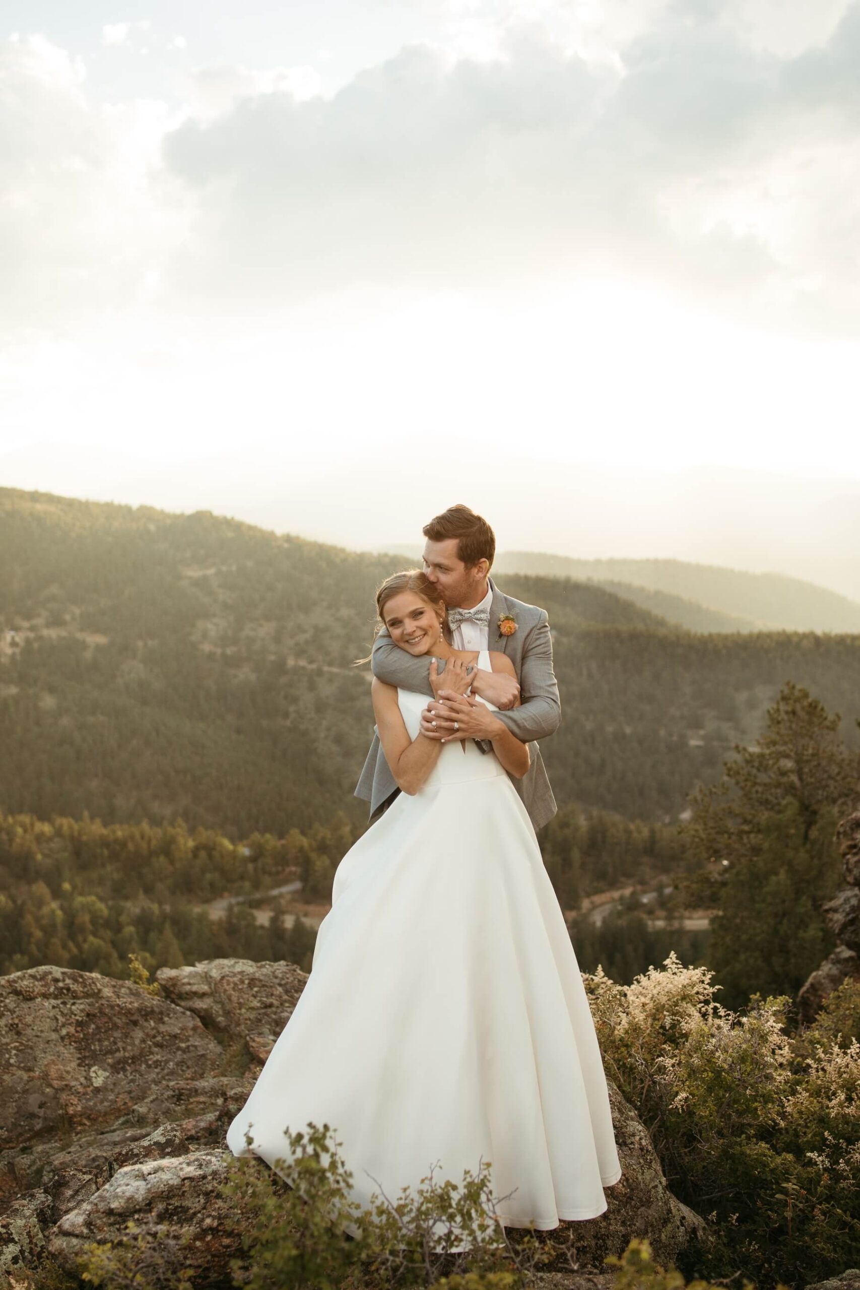 Groom standing behind bride kissing her head with mountains in the background