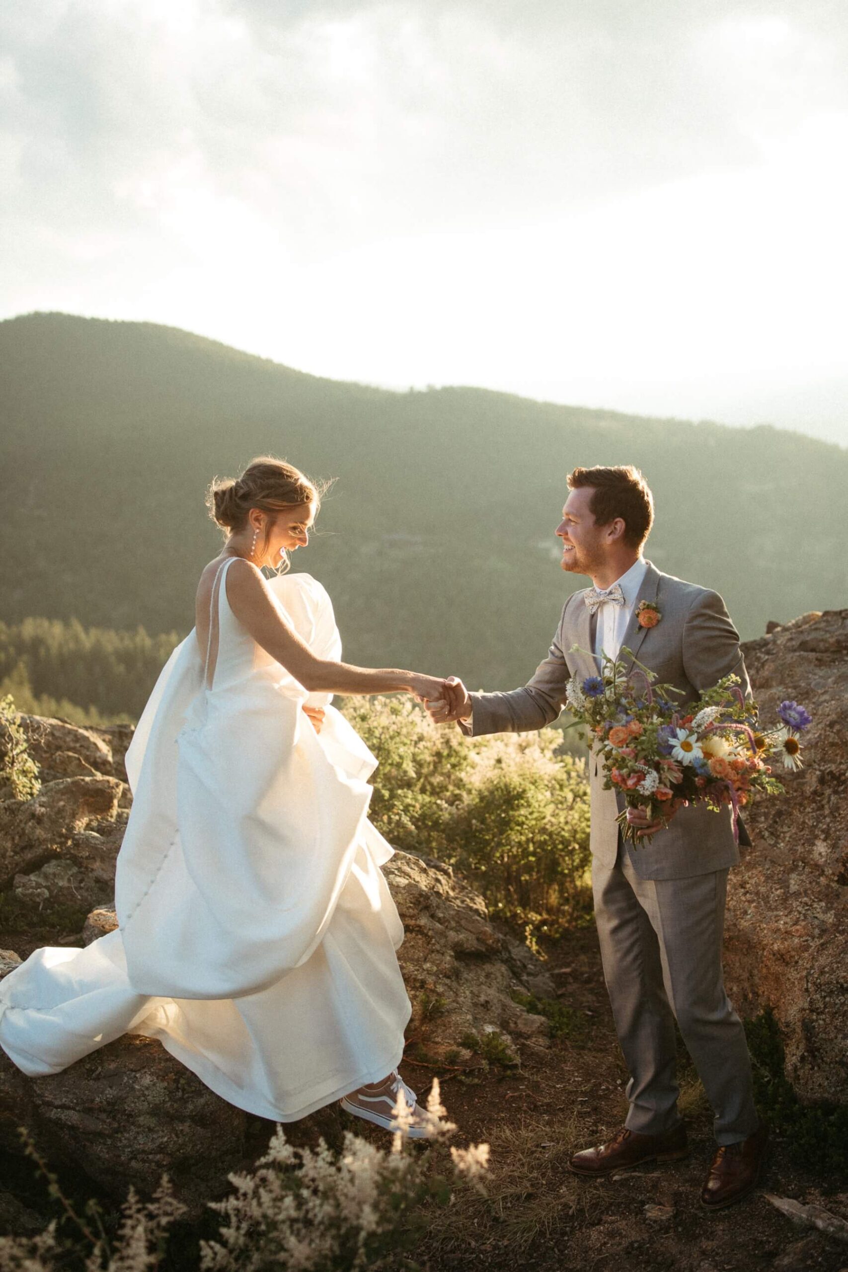 Groom helping bride climb down from rocks while holding her bouquet 