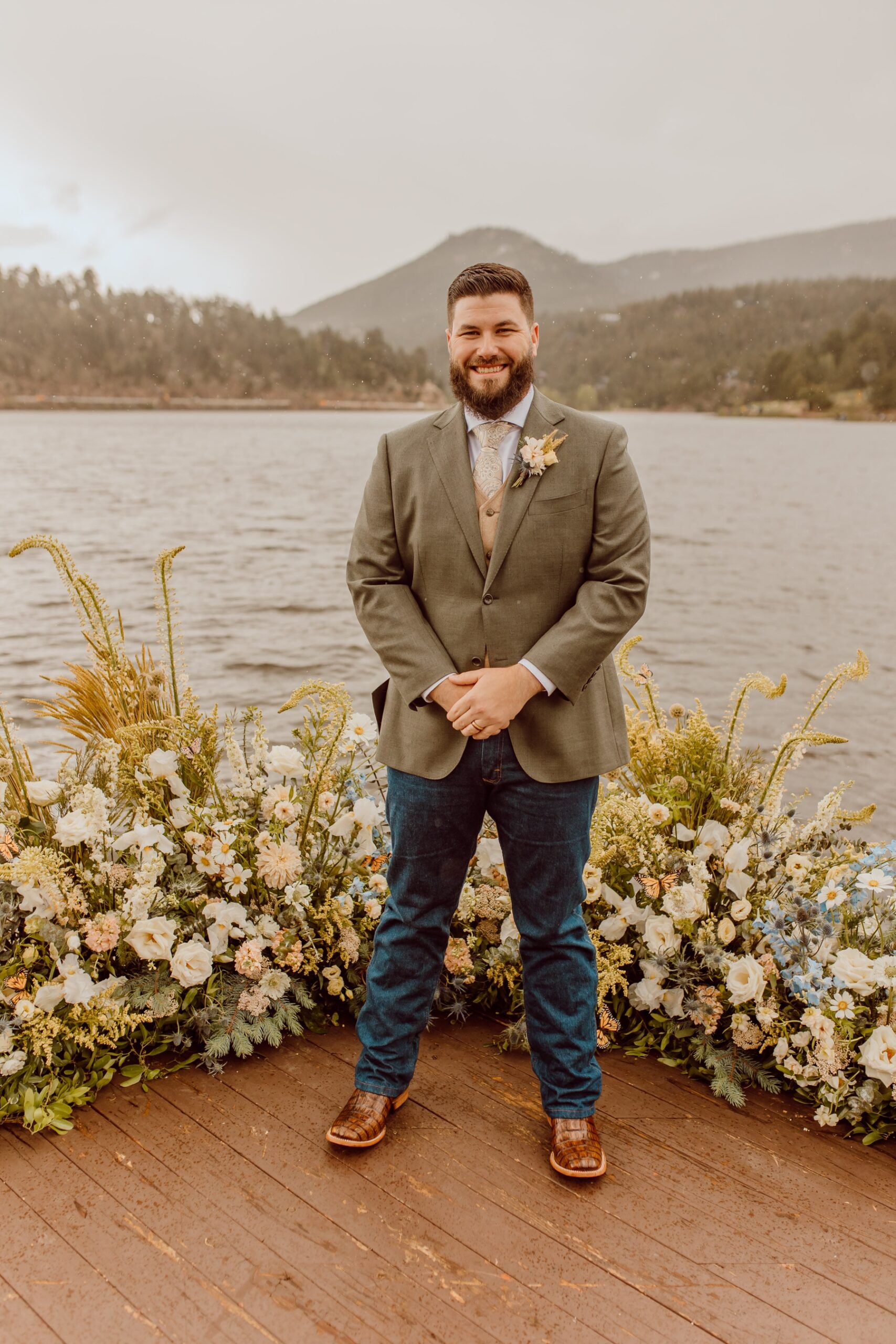 Groom in vest and suit jacket with boots and jeans standing on deck of Evergreen Lake House
